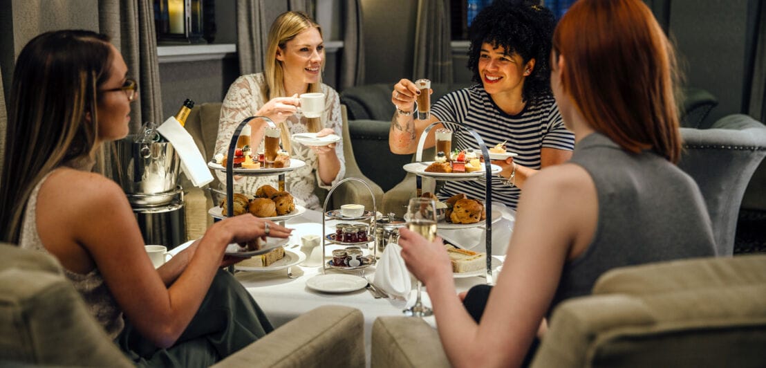 Vier Frauen an einem edel gedeckten Tisch bei einer Teatime mit Gebäck und Champagner