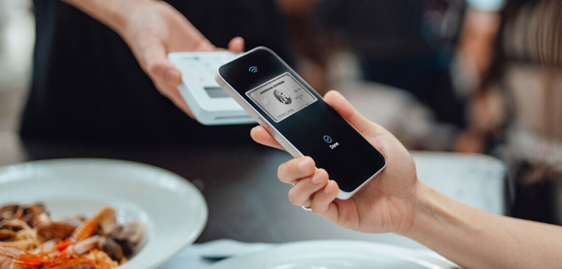 Kartenzahlung mit einer Amex Platinum Card per Smartphone an einem Kartenlesegerät in einem Restaurant.