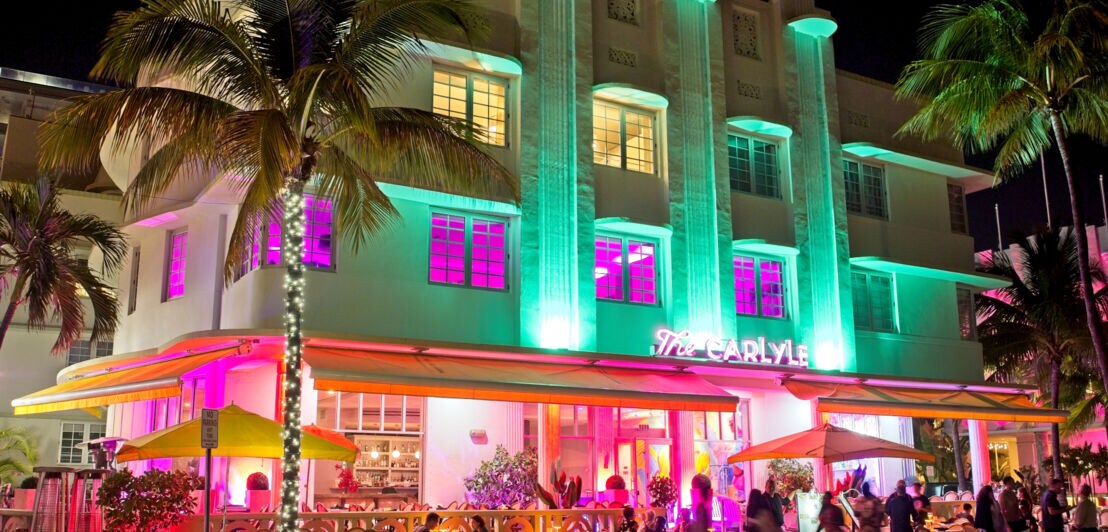 In Neonfarben beleuchtetes Hotel im Art-Deco-Stil mit Außengastronomie und Personen auf dem Gehweg bei Nacht.