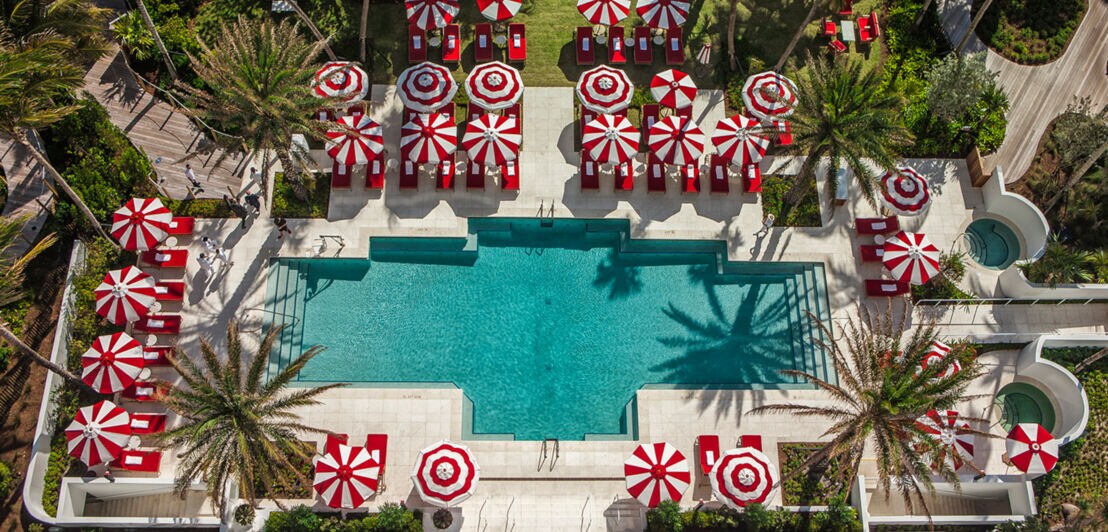 Palmengesäumter Hotelpool mit Liegen und rot-weiß-gestreiften Sonnenschirmen aus der Vogelperspektive.
