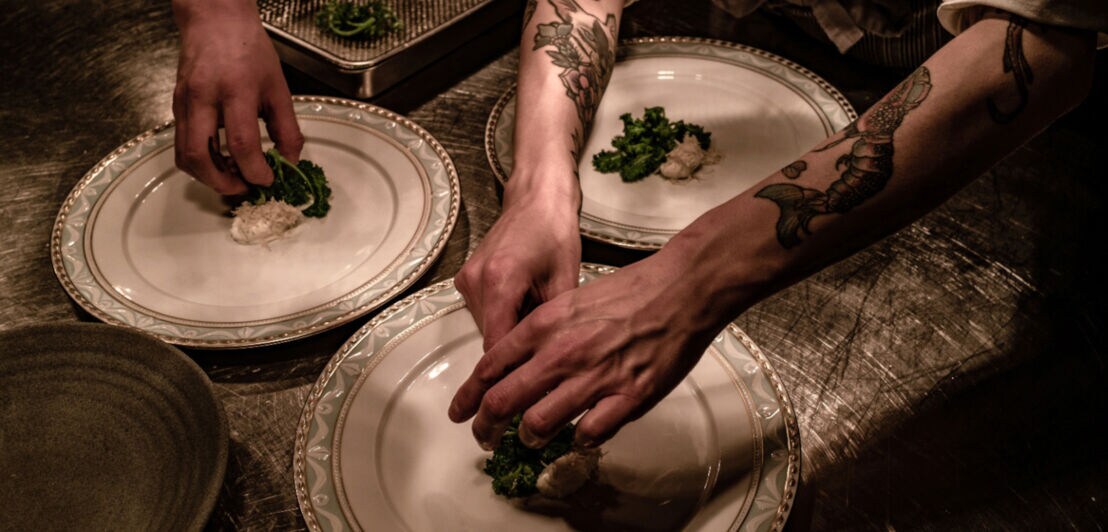 Nahaufnahme von Händen beim Anrichten von Speisen auf großen, weißen Tellern in einer Restaurantküche.