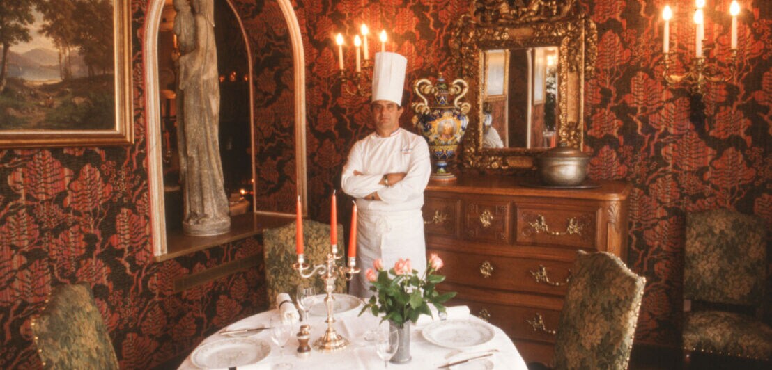 Koch Paul Bocuse vor einem edel gedeckten Tisch im Innenraum eines opulent dekorierten Restaurants mit floralem Wandmuster