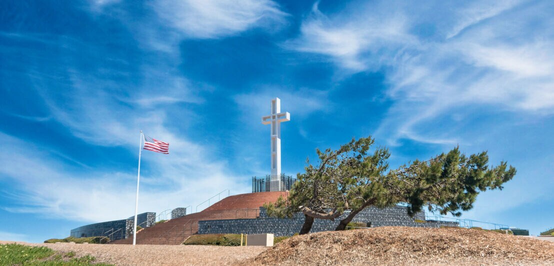 Eine Gedenkstätte mit einem weißen Kreuz auf einem Hügel, daneben ein Mast mit der US-amerikanischen Flagge