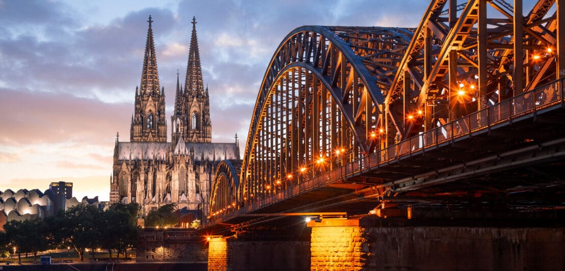 Hinter einer Brücke thront der angeleuchtete Kölner Dom in der Dämmerung