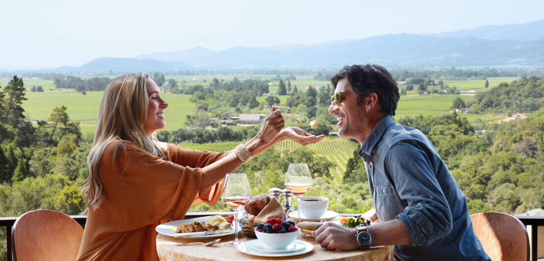  Ein Paar sitzt auf einer Terrasse beim Frühstück vor grünem Landschaftspanorama, sie reicht ihm einen Probierbissen auf der Gabel über den Tisch.