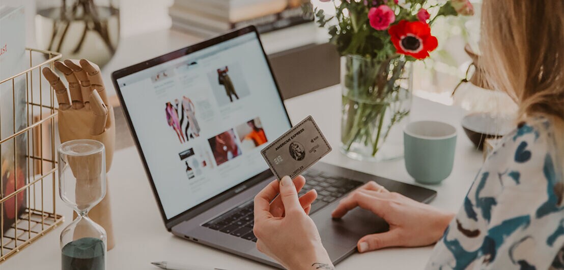Eine Platinum Kreditkarte von American Express in der Hand einer Frau beim Onlineshopping an einem Laptop auf einem Schreibtisch.