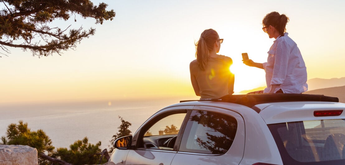 Rückansicht zweier junger Frauen auf dem Dach eines weißen Autos an einer Meeresküste bei Sonnenuntergang.