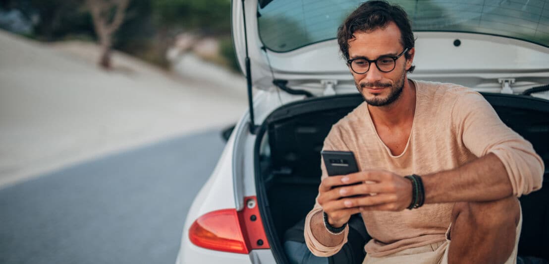 Ein mittelalter Mann in Freizeitkleidung sitzt im geöffneten Kofferraum eines Autos und bedient lächelnd sein Smartphone.