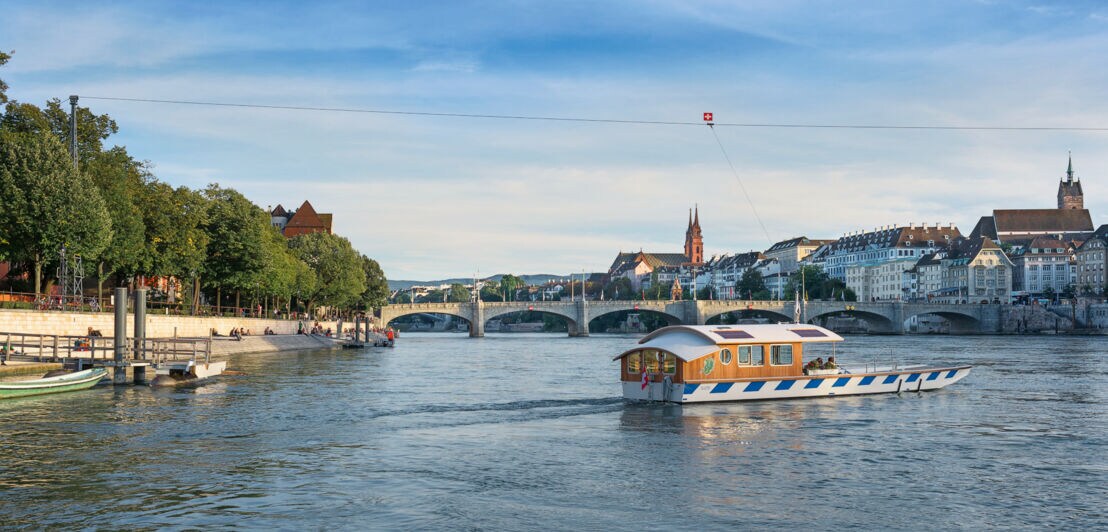 Eine kleine Fähre fährt an einem Seil entlang über den Rhein, im Hintergrund die Basler Altstadt.