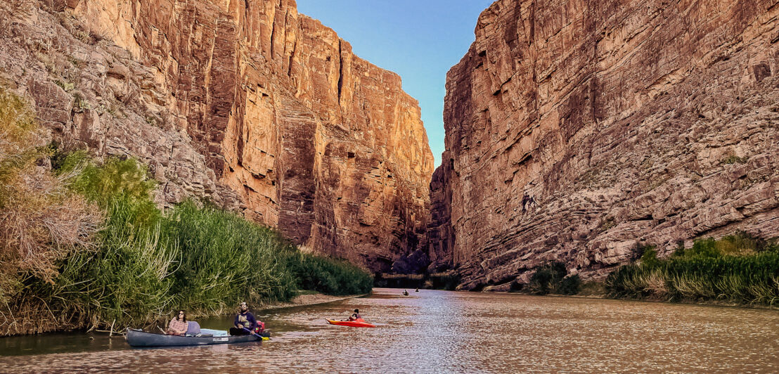 Personen in Kayaks auf einem Fluss in einem Canyon mit rötlichen Felsen.