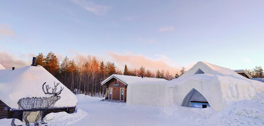 Mehrere Gebäude in einer Schneelandschaft und ein aus Ästen gebauter Hirsch.