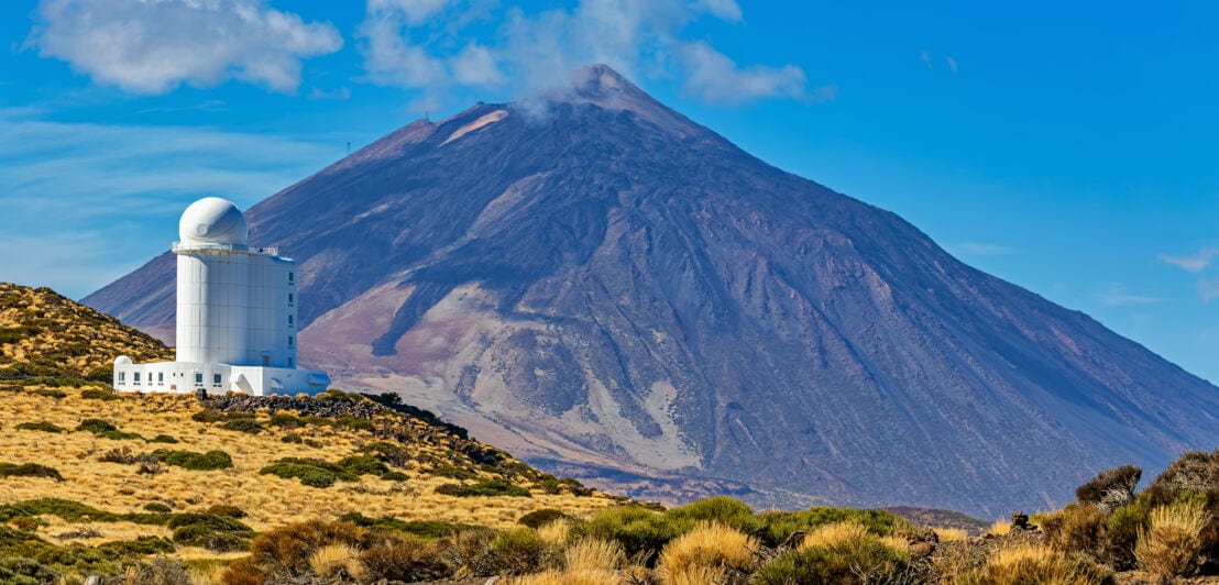 Ein weißes Observatorium in karger Landschaft, im Hintergrund ein Vulkan.