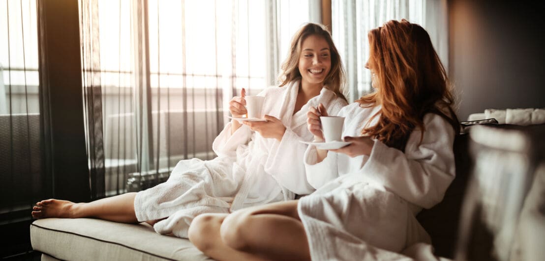 Zwei fröhliche, junge Frauen in weißen Bademänteln sitzen mit Kaffeetassen auf einem Sofa.