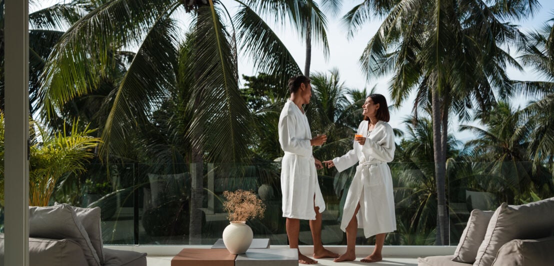 Ein Paar in weißen Bademänteln steht auf einer luxuriösen Terrasse vor einer Palmenlandschaft.