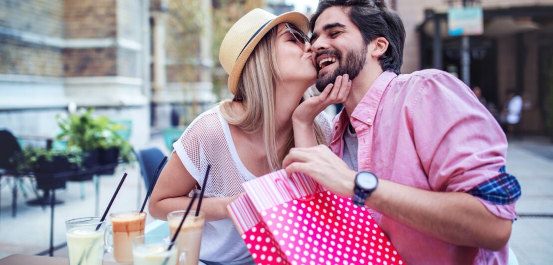 Ein glückliches Paar mit bunter Geschenktüte an einem Tisch in einem Straßencafé, sie gibt ihm einen Kuss auf die Wange.