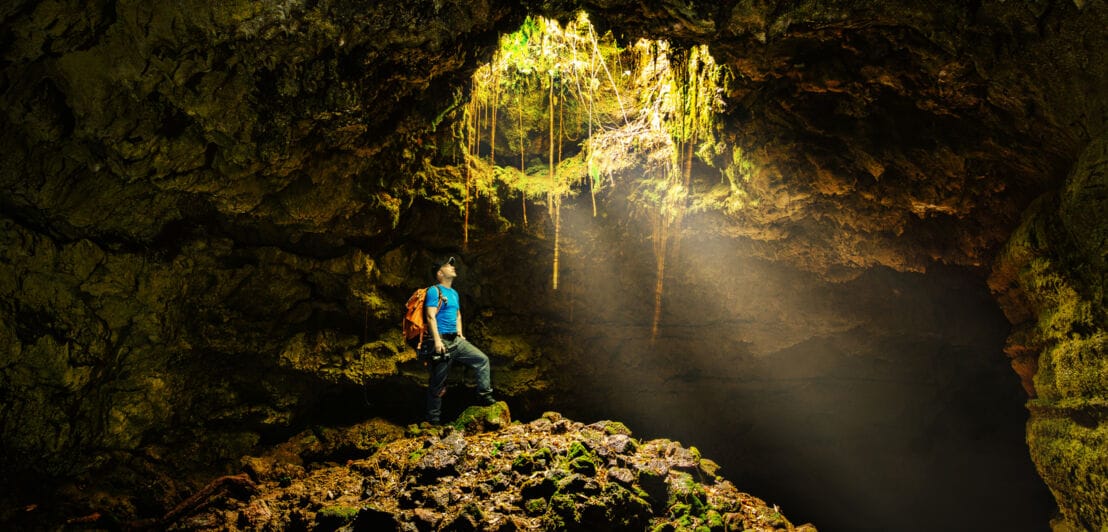 Ein Mann in einer Felshöhle schaut durch ein Loch über ihm ins Licht.