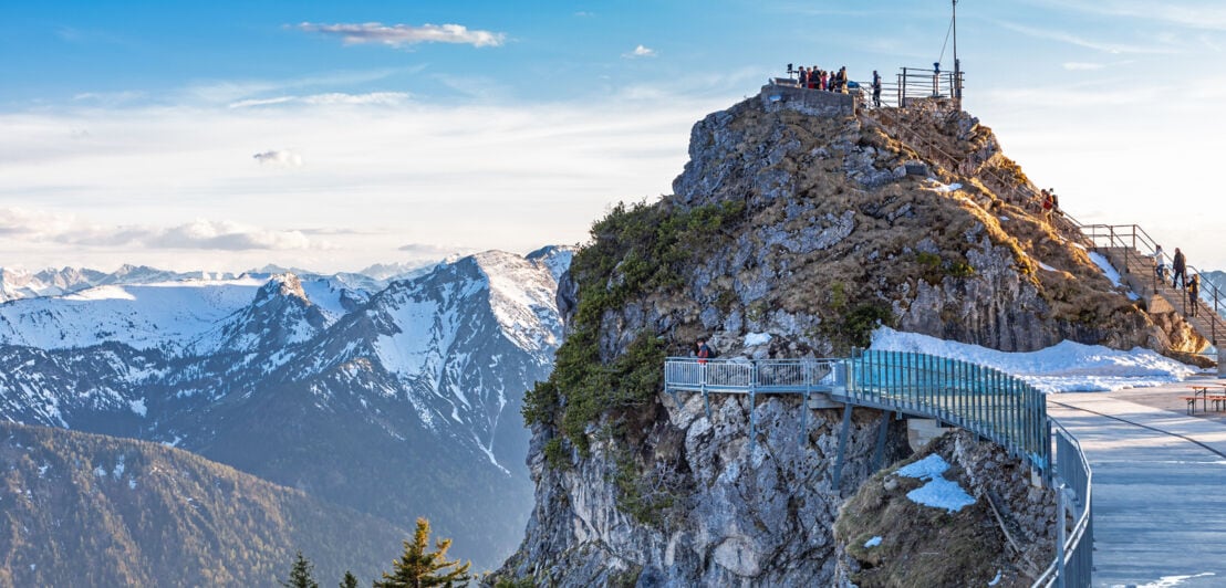 Personen auf einer Aussichtsplattform auf einem Berggipfel vor verschneitem Alpenpanorama.
