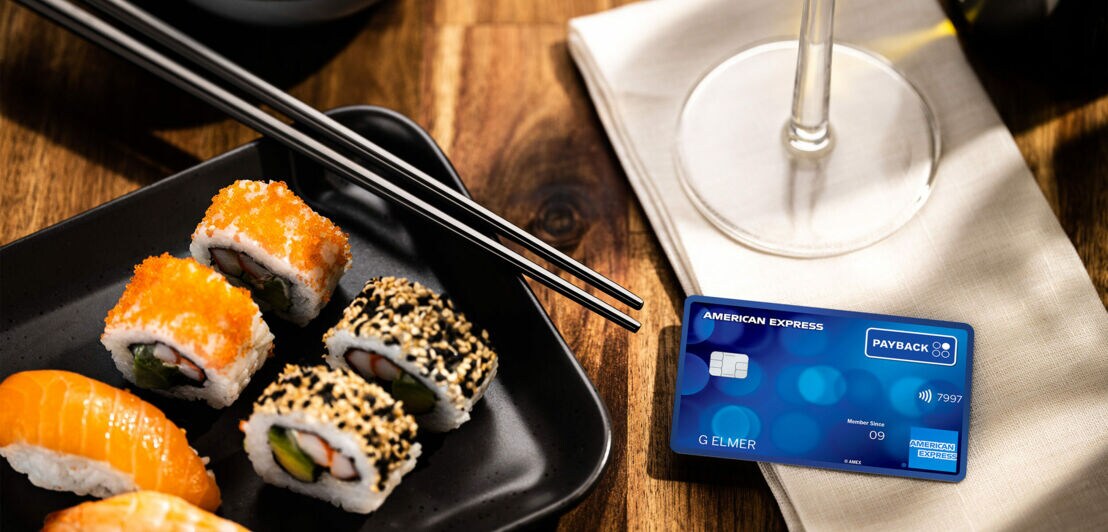 Eine PAYBACK American Express Kreditkarte liegt auf einem Tisch auf einer Stoffserviette neben einem Teller mit Sushirollen in einem Restaurant