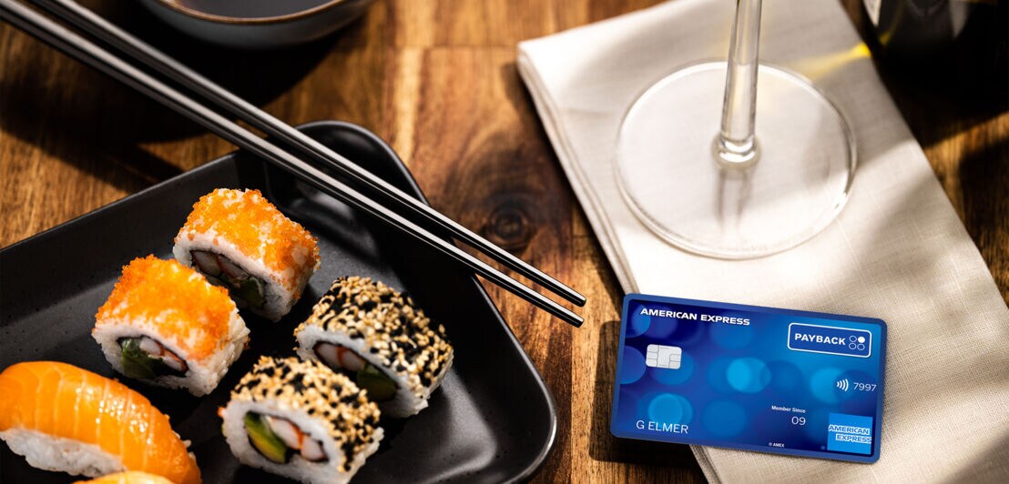 Eine PAYBACK American Express Kreditkarte liegt auf einem Tisch auf einer Stoffserviette neben einem Teller mit Sushirollen in einem Restaurant