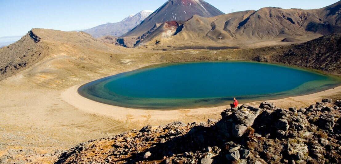 Eine Person sitzt auf einem Felsen vor einem Bergsee inmitten einer Vulkanlandschaft.