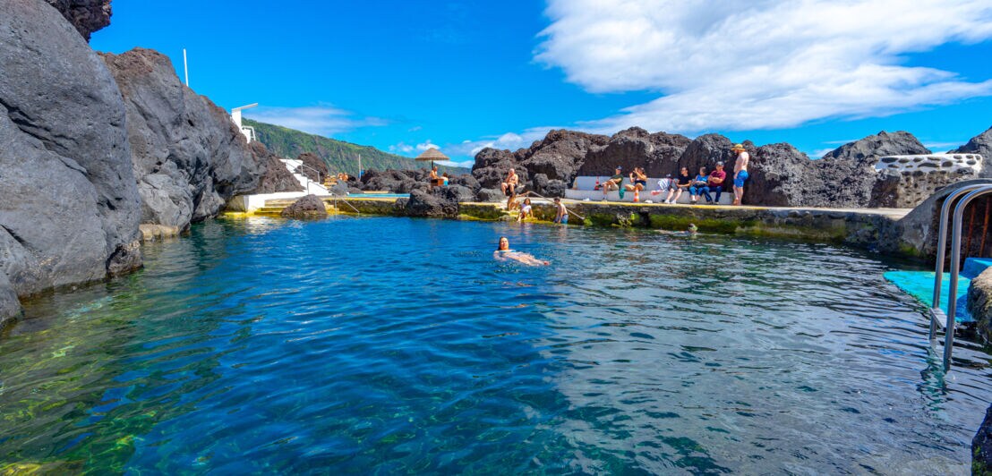Personen an und in einem Naturschwimmbecken, umgeben von Felsbrocken.