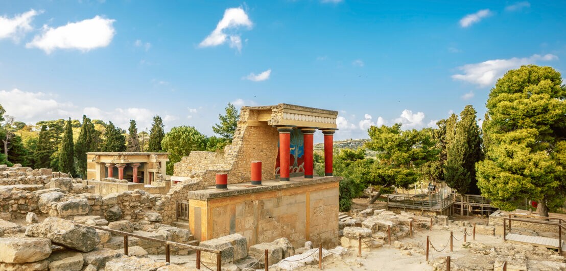 Ruinenstätte des Palastes von Knossos mit roten Säulen, umgeben von Bäumen.