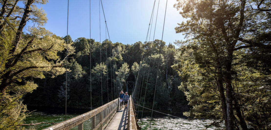 Zwei Personen laufen auf einer Hängebrücke über einen Fluss in einem Waldgebiet.