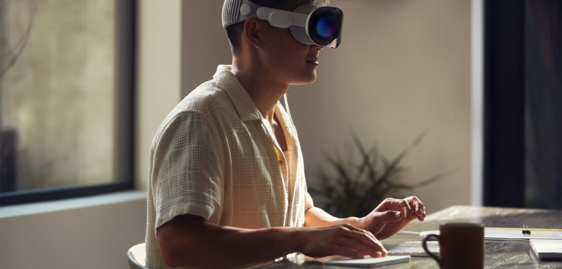 Eine Person sitzt mit einer Apple VR-Brille an einem Schreibtisch.
