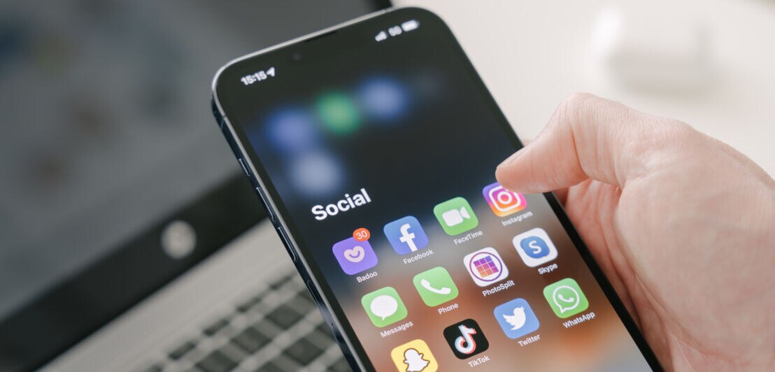 Eine Hand hält ein iPhone mit Social-Media-Apps auf dem Display vor dem Hintergrund eines Laptops.