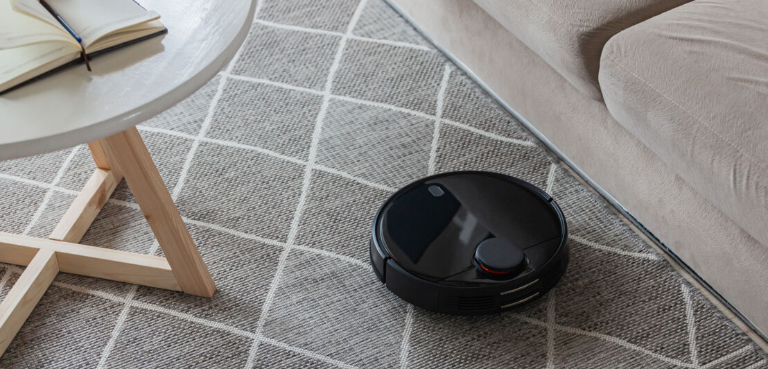 Ein schwarzer Saugroboter auf einem Teppich vor einem Sofa.