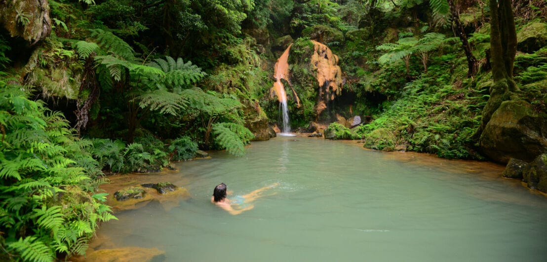 Eine Person badet in einem Teich vor einem Wasserfall inmitten tropischer Natur.