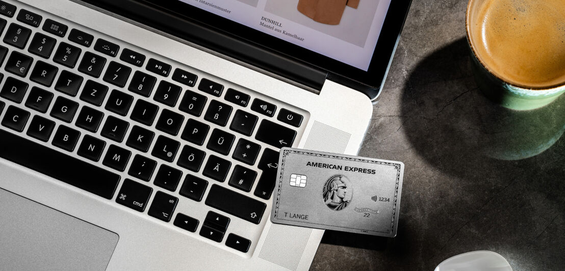 Aufsicht einer silbernen Kreditkarte von American Express auf einem Laptop neben einer Tasse Kaffee auf einem Tisch.