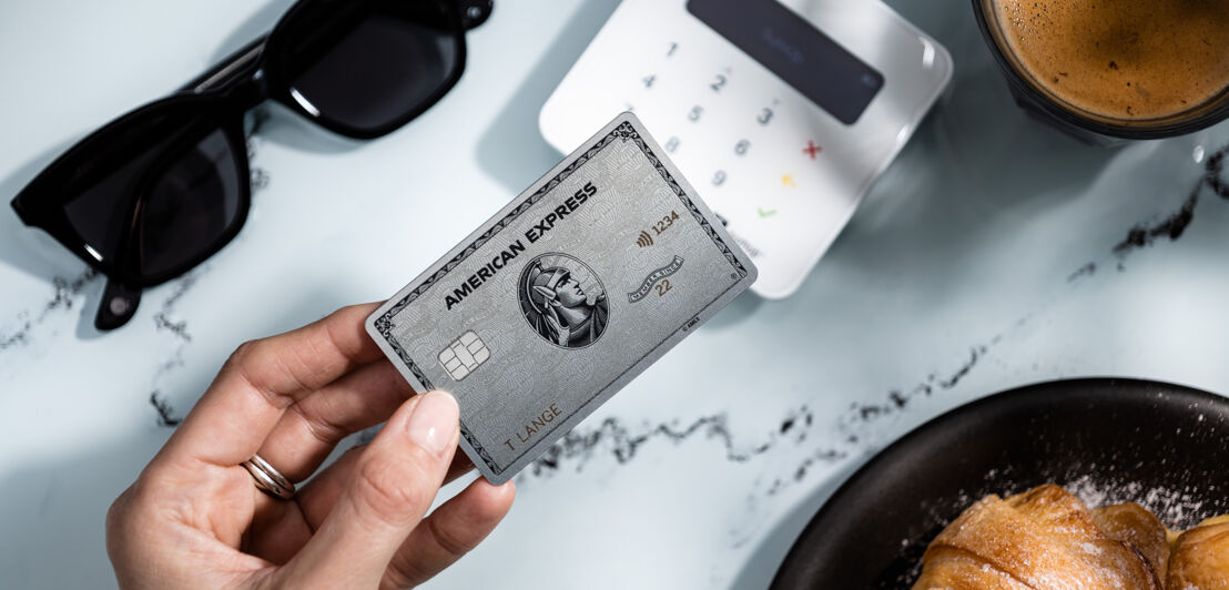 Kontaktlose Bezahlung mit einer Amex Platinum Card an einem Kartenlesegerät auf einem Tisch neben Kaffee, Gebäck und Sonnenbrille.