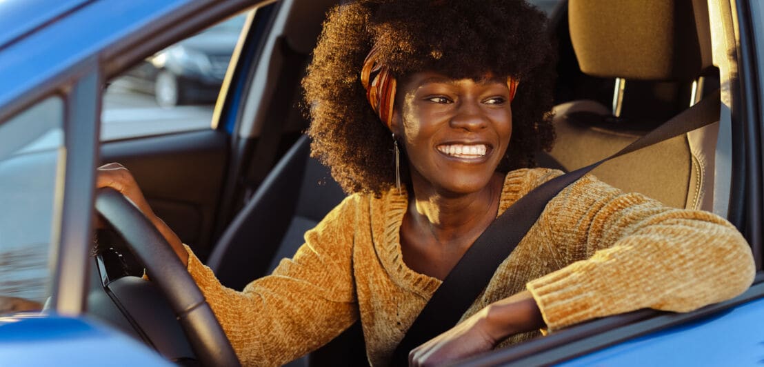 Eine lachende Frau am Steuer eines blauen Autos bei geöffnetem Fenster.