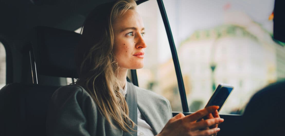 Eine junge Frau auf dem Rücksitz eines Autos mit Smartphone in den Händen blickt aus dem Fenster.