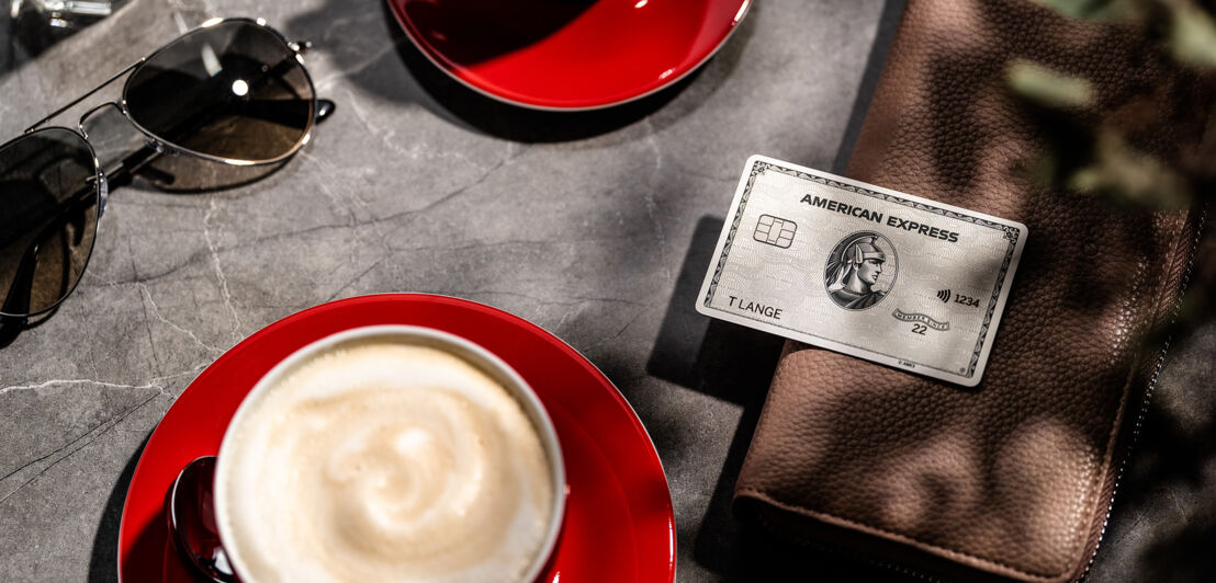 Eine Amex Platinum Card liegt auf einer Geldbörse auf einem Tisch neben einer roten Tasse mit Milchkaffee im Sonnenschein