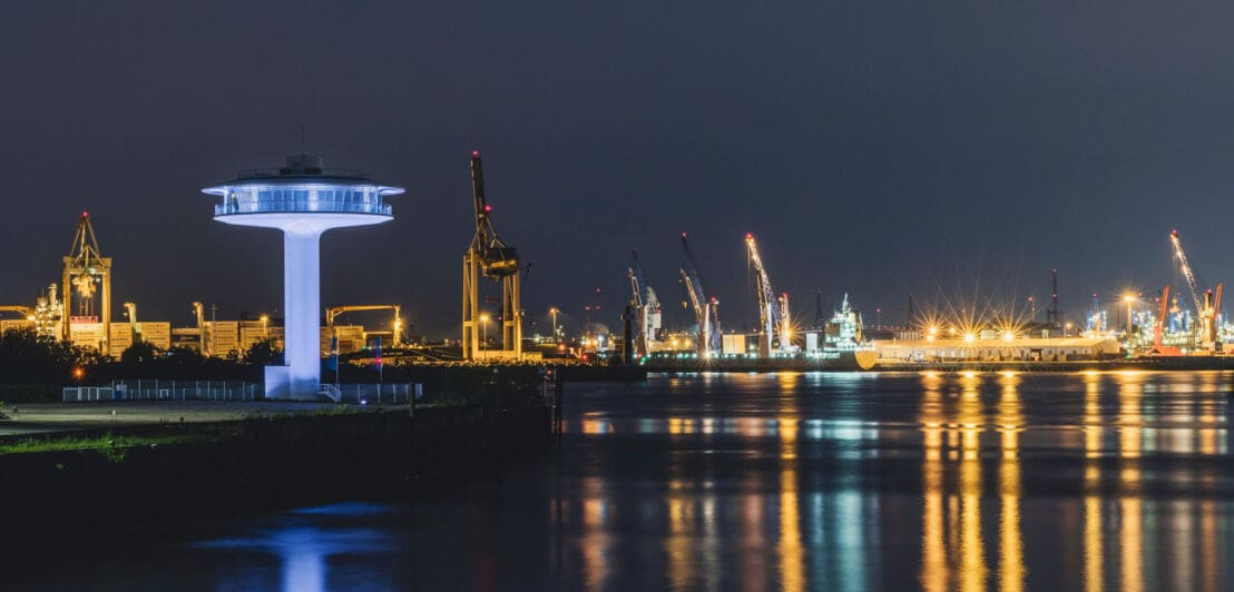 Beleuchteter Leuchtturm in Ufo-Form mit Panoramafenstern vor der Skyline des beleuchteten Hamburger Hafens bei Nacht.