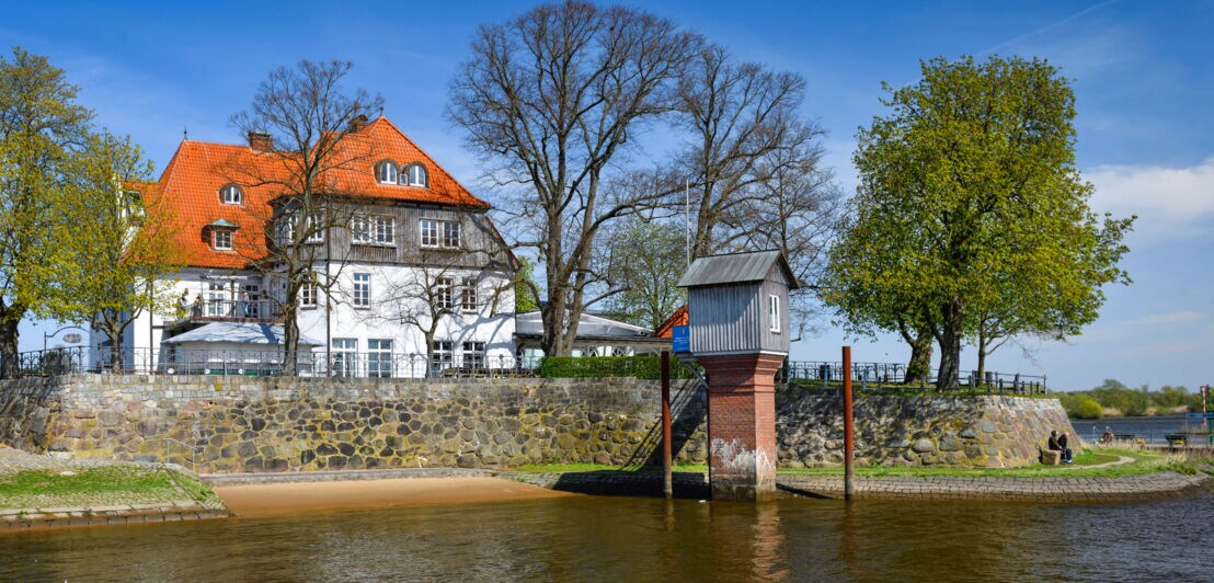 Ein Pegelhaus im Wasser vor einer Uferpromenade mit Fährhaus in begrünter Umgebung.