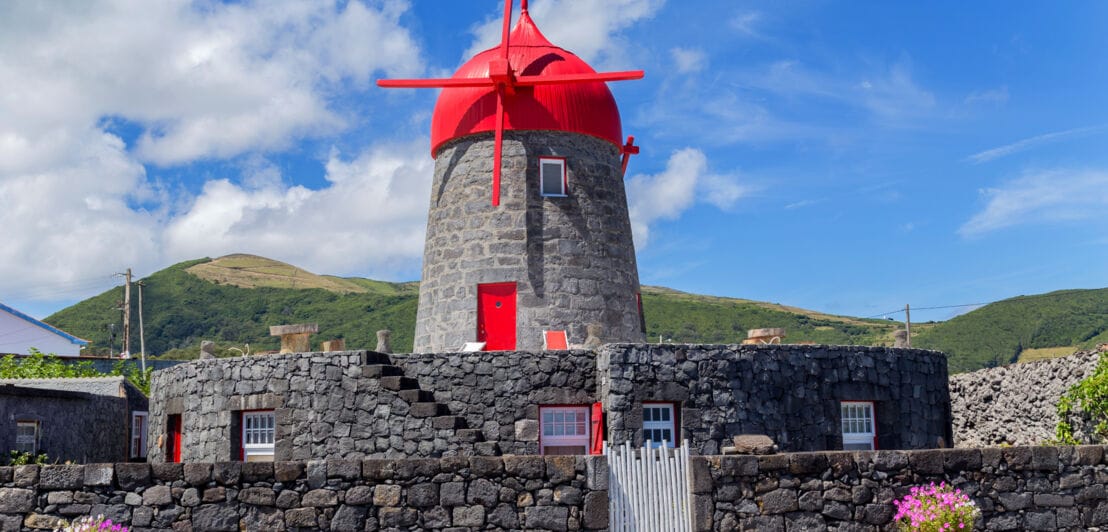 Windmühle aus Stein mit roter Kuppel, umgeben von einer Steinmauer vor grüner Hügellandschaft.