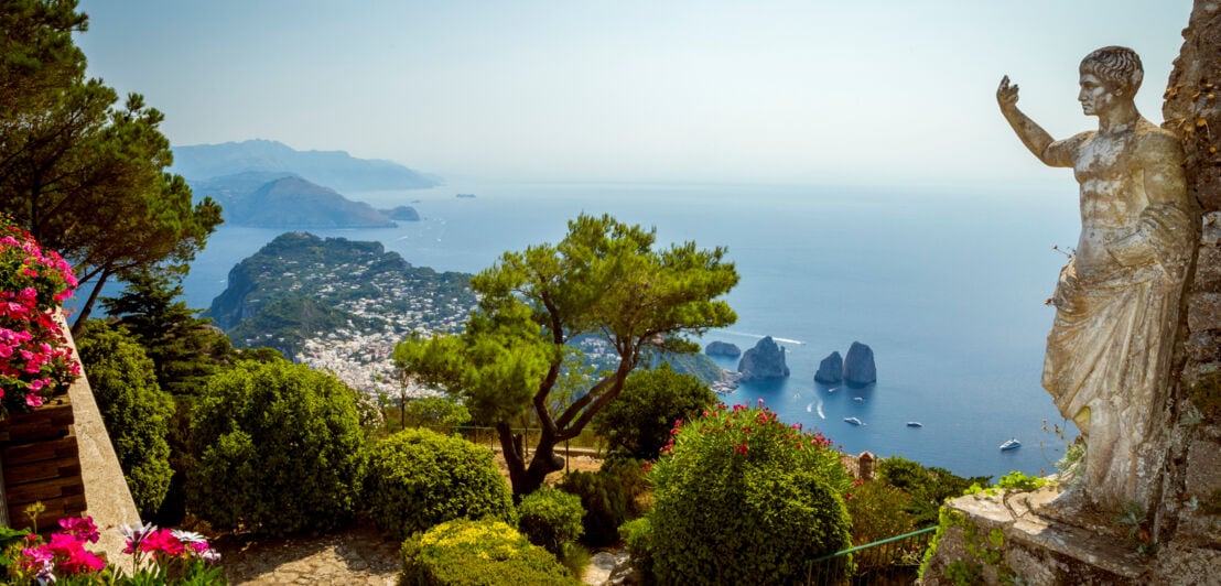 Blick von einer mediterranen Parkanlage mit Steinstatue auf einem Berg hinunter auf eine Insellandschaft mit Felsformationen im Meer.