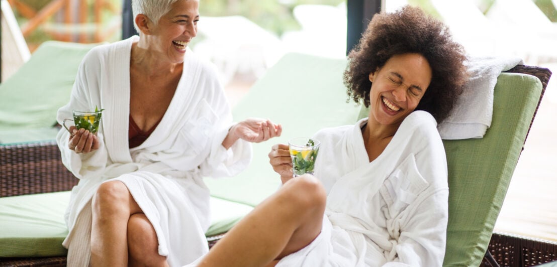 Zwei gut gelaunte Frauen in weißen Bademänteln mit Teegläsern in den Händen auf Liegen in einem Wellnessbereich.