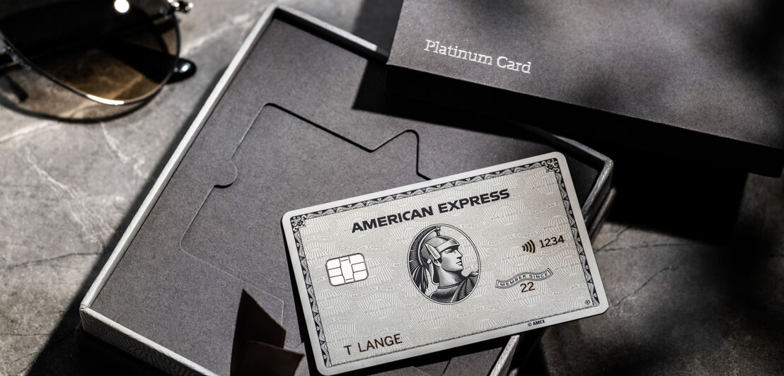 Eine American Express Platinum Card aus Metall liegt auf einer geöffneten Präsentationsbox aus grauer Feinkartonage mit Platinum Card Aufschrift auf einem Marmortisch