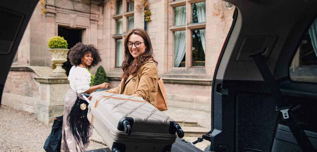 Blick durch den geöffneten Kofferraum eines Autos auf zwei junge Frauen mit Gepäck vor einem Hoteleigang, eine Frau nimmt einen Koffer von der Ladefläche.