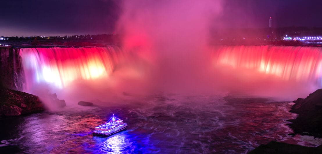 Ein blau beleuchtetes Ausflugsboot vor den rot beleuchteten Niagarafällen bei Nacht.