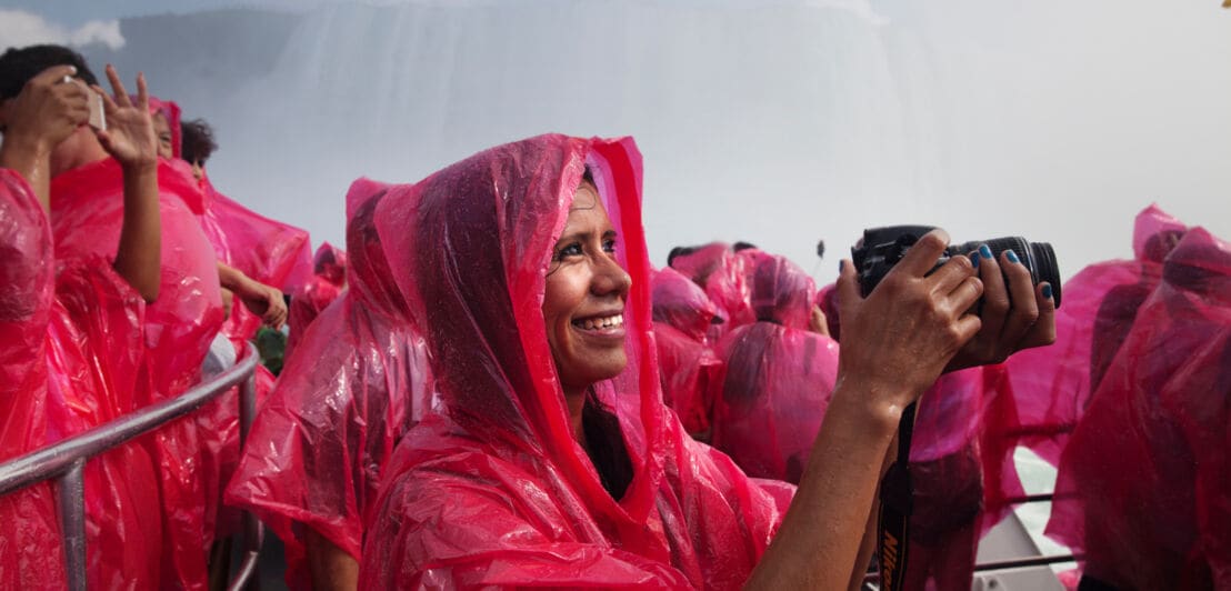 Nahaufnahme einer Frau im roten Regencape mit einer Kamera in ihren Händen auf einem Ausflugsboot vor einem Wasserfall.