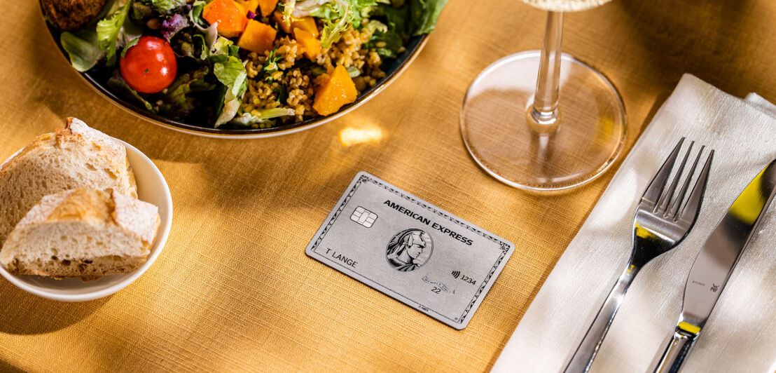 Eine silberne Kreditkarte von American Express liegt auf einer gelben Tischdecke neben einem Salatteller auf einem Restauranttisch.