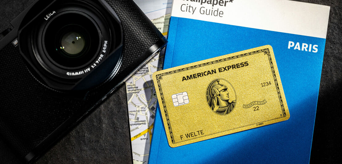 Aufsicht einer goldenen Kreditkarte von American Express auf einem Reiseführer mit Stadtplan neben einer Kamera.