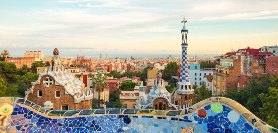 kyline von Barcelona am Meer, im Vordergrund eine geschwungene Mauer mit Mosaiksteinen vor zwei weiteren Bauwerken von Gaudí.