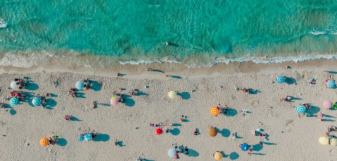 Belebter, weißer Sandstrand mit Sonnenschirmen am türkisblauen Wasser aus der Vogelperspektive.