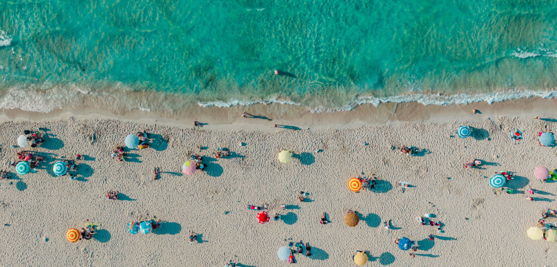 Belebter, weißer Sandstrand mit Sonnenschirmen am türkisblauen Wasser aus der Vogelperspektive.